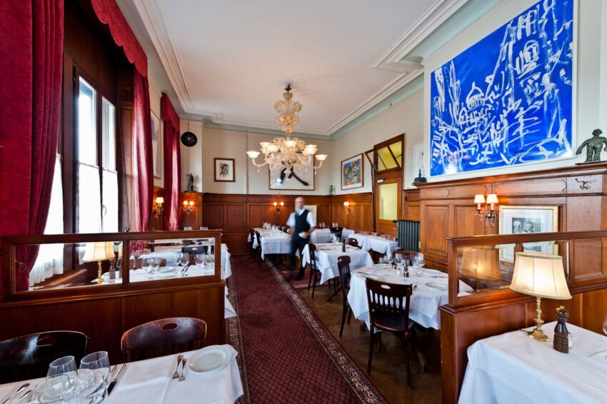 Elegant restaurant interior with white tablecloths, chandelier, and red curtains, waiter setting tables in a cozy atmosphere.