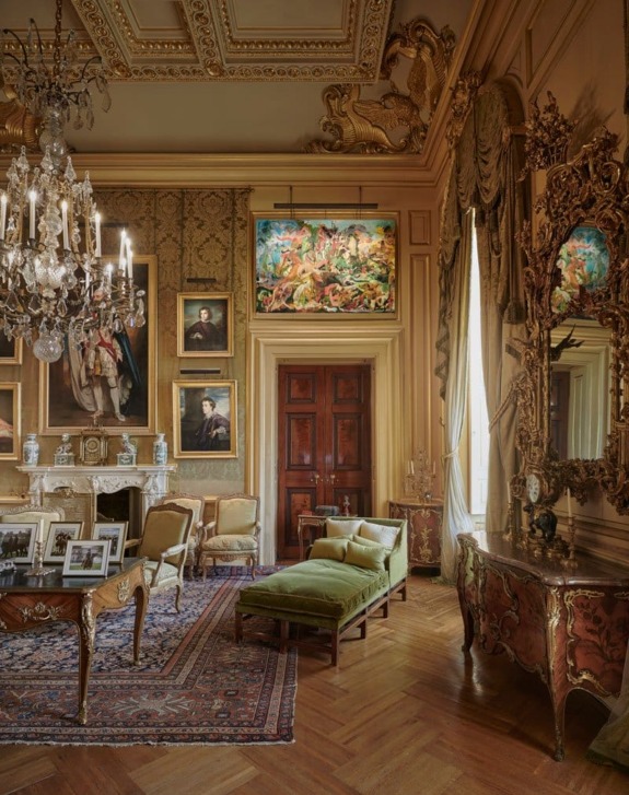Elegant room with ornate chandelier, paintings, vintage furniture, and decorative molding on walls and ceiling.