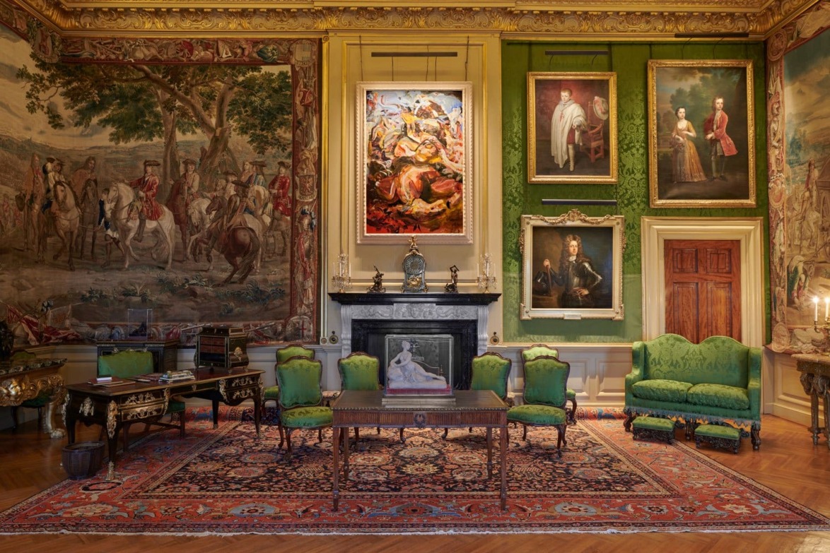 Luxurious ornate room with green walls, vintage furniture, elaborate tapestry, and classical paintings above a fireplace.