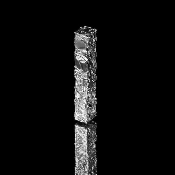 Tall textured silver sculpture standing against a black background with a reflective surface.