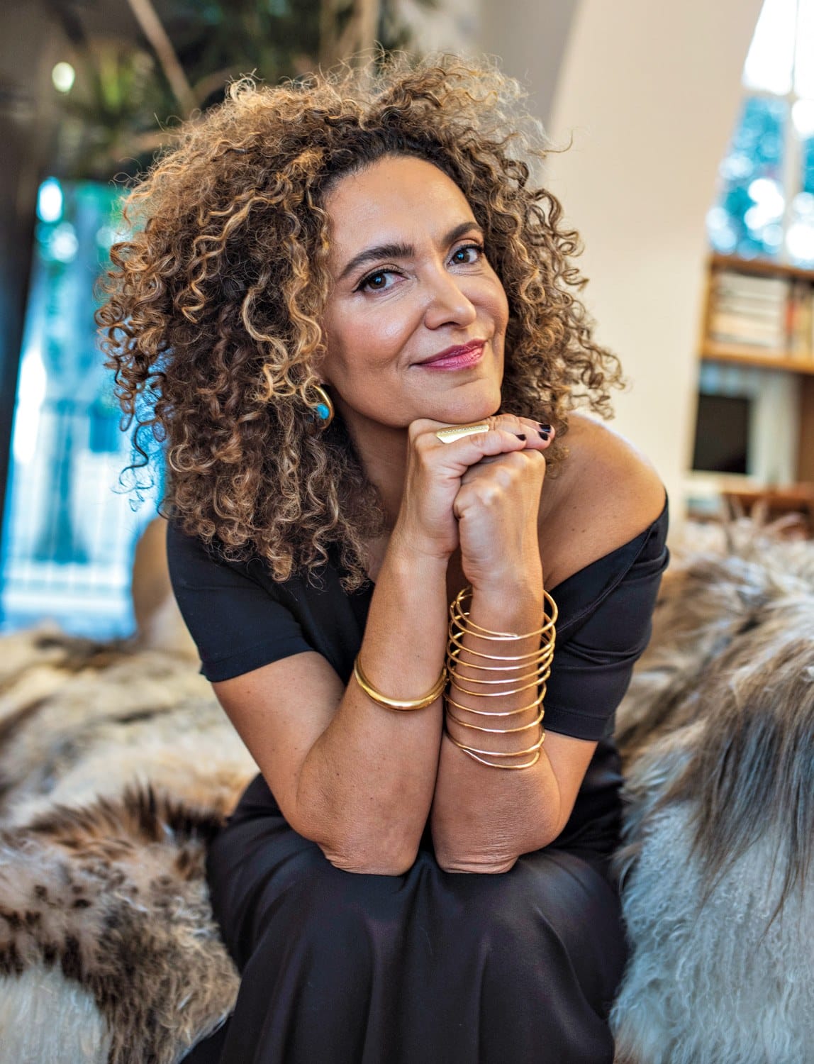 Person sitting on a fur-covered chair, wearing a black dress and gold bangles, with curly hair and a thoughtful expression.