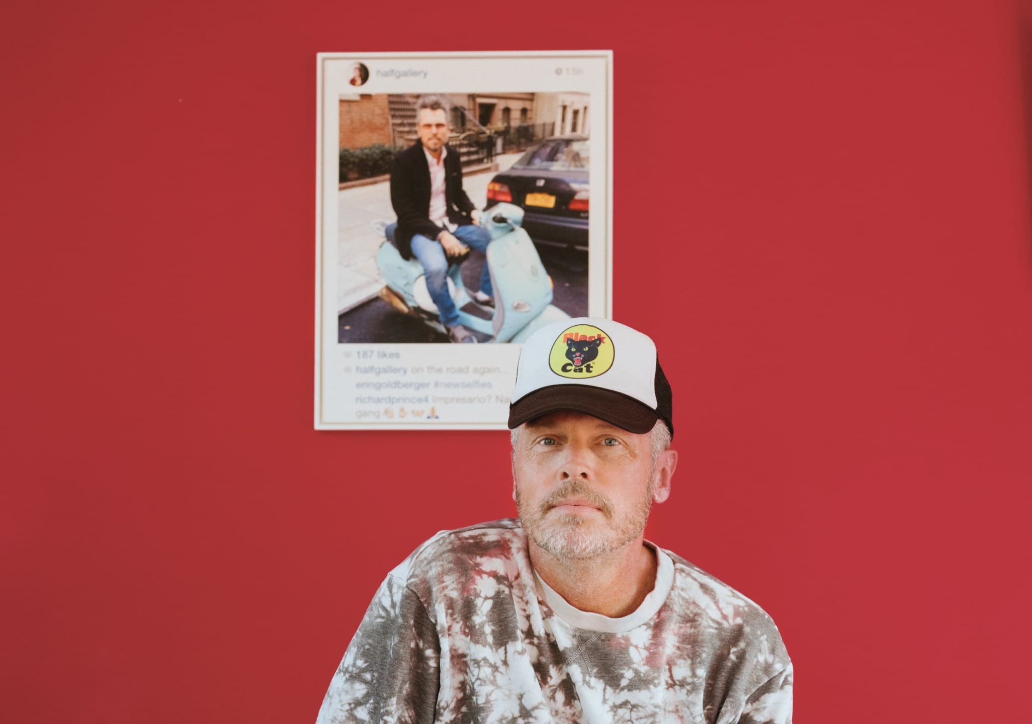 Man wearing a cap and tie-dye shirt sitting in front of a red wall with a framed photo behind him.