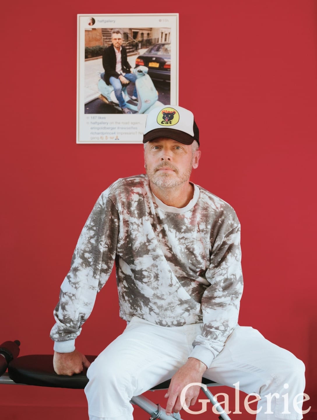 Man in tie-dye shirt and baseball cap sits with red wall and framed photo background, "Galerie" text at bottom right.