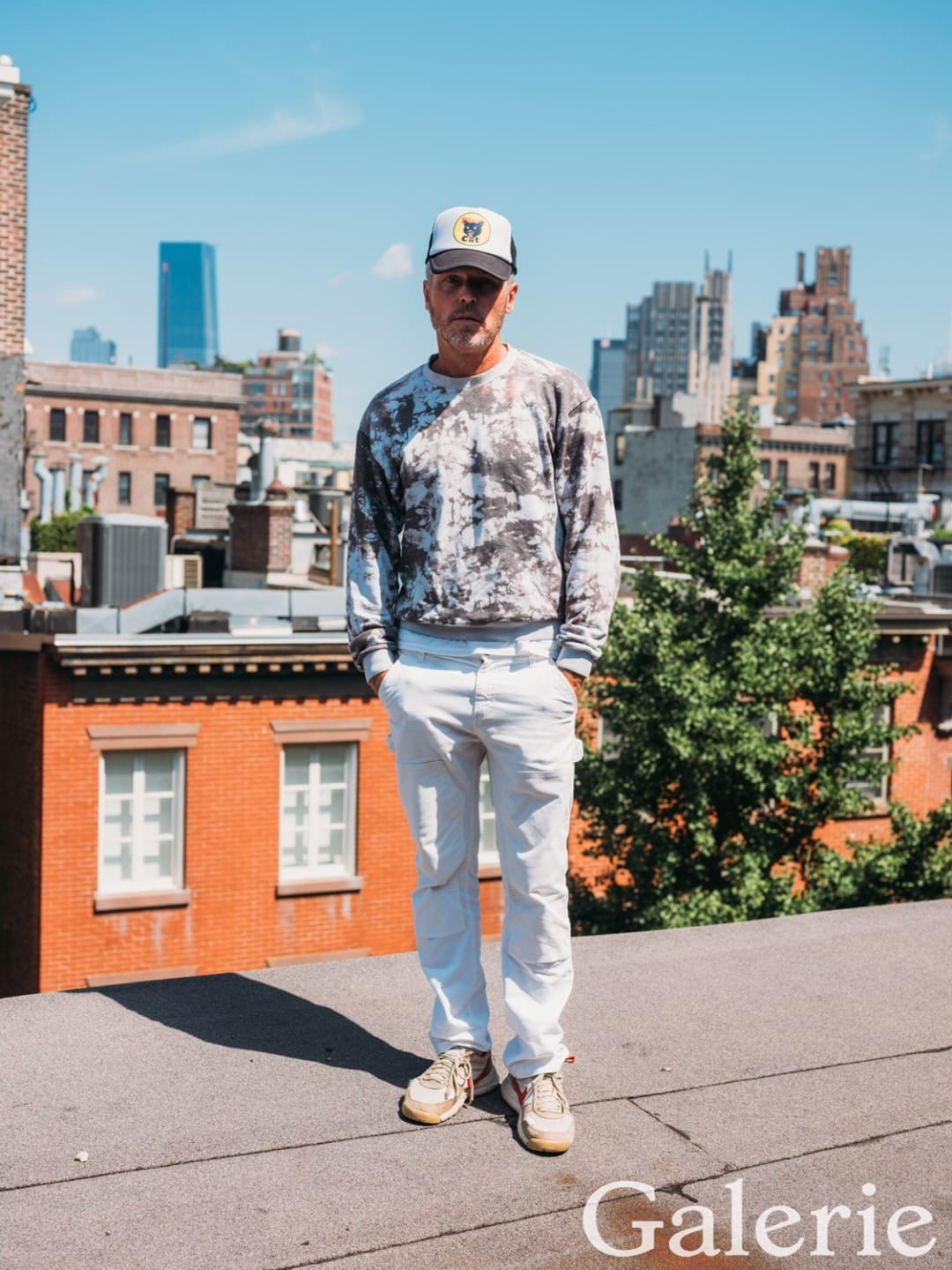 Man in casual attire standing on rooftop with city buildings in background, wearing a cap and patterned sweater.
