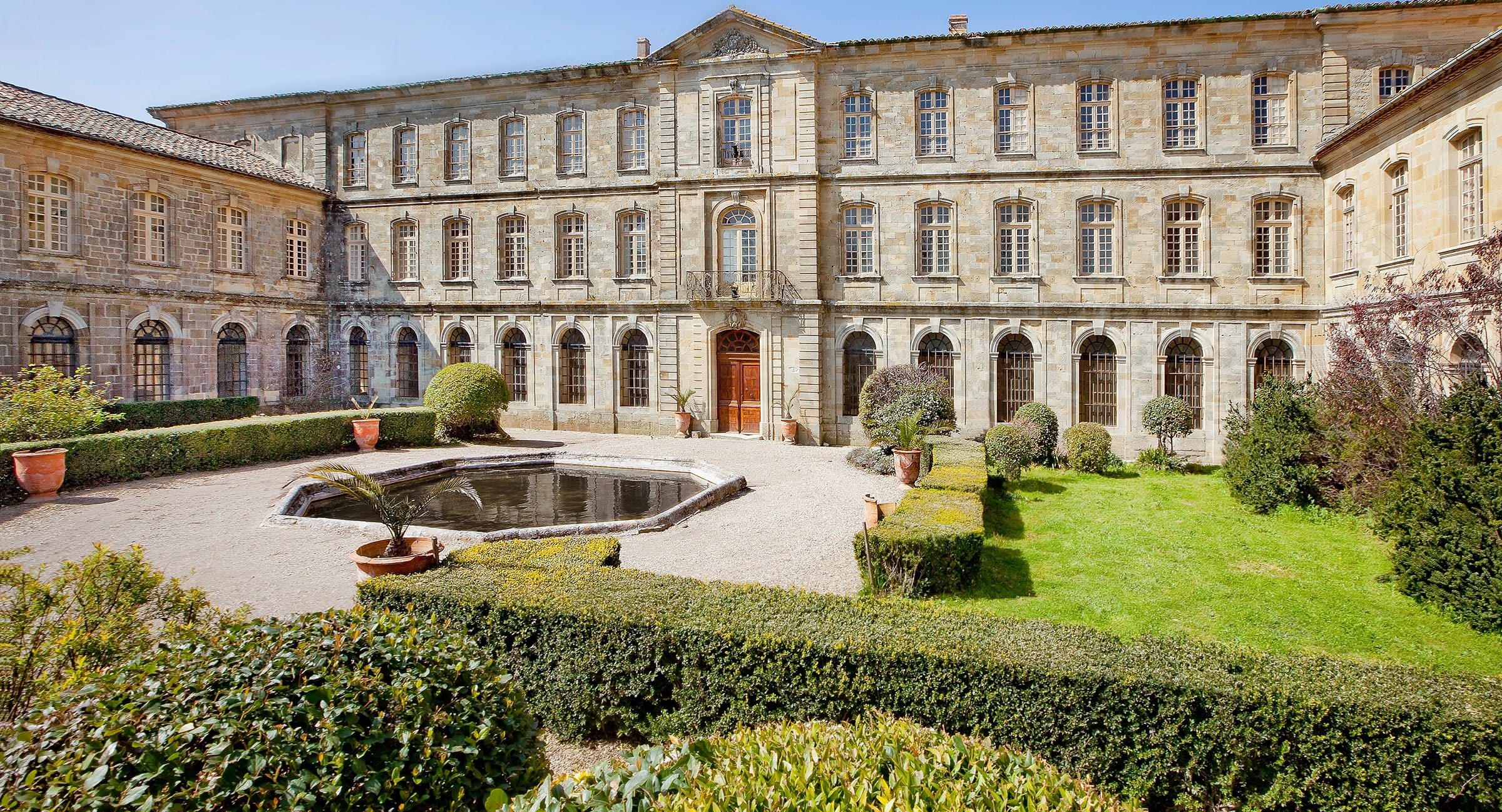 Historic stone building with arched windows, courtyard garden, and small pond in foreground.