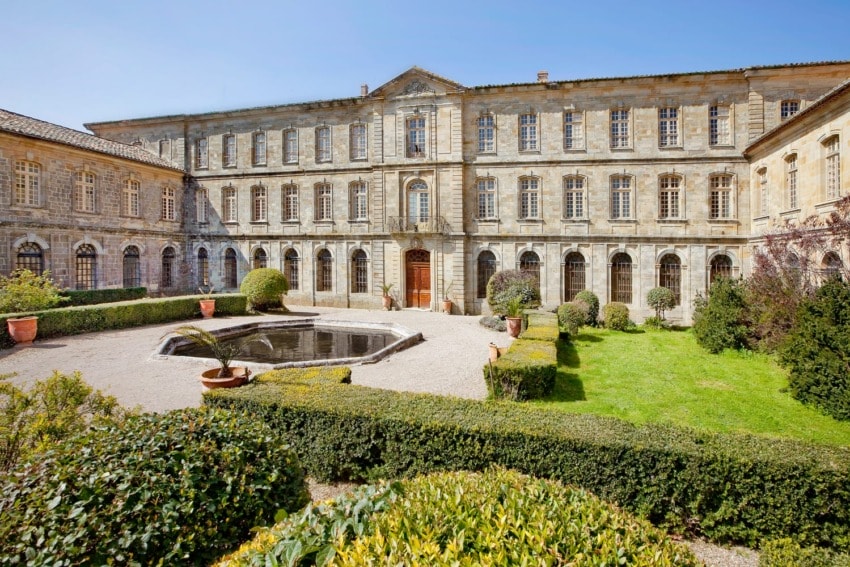 Historic stone building with a courtyard, fountain, and trimmed hedges on a sunny day.