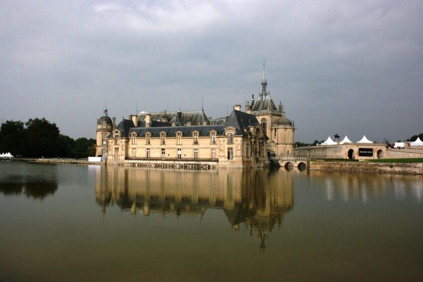 Historic château reflected in a calm lake under a cloudy sky, with white tents visible in the background.