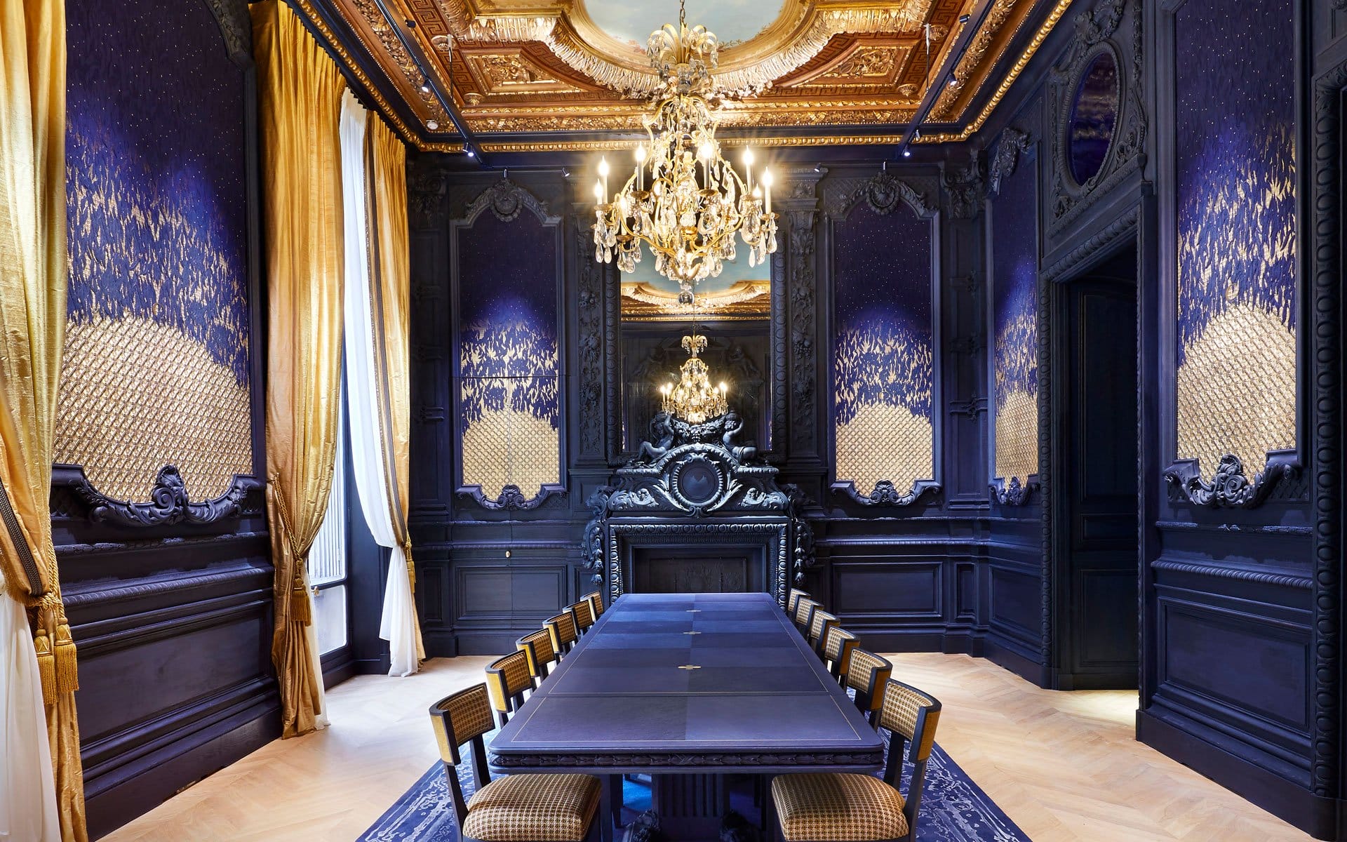 Luxurious dining room with ornate blue and gold decor, a long table, elegant chairs, and a grand chandelier.