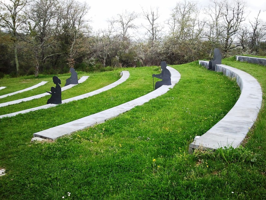 Outdoor amphitheater with metal silhouette sculptures seated on stone benches among grass and trees in the background.