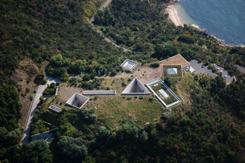 Aerial view of hilltop art museum with geometric structures surrounded by greenery near a shoreline.
