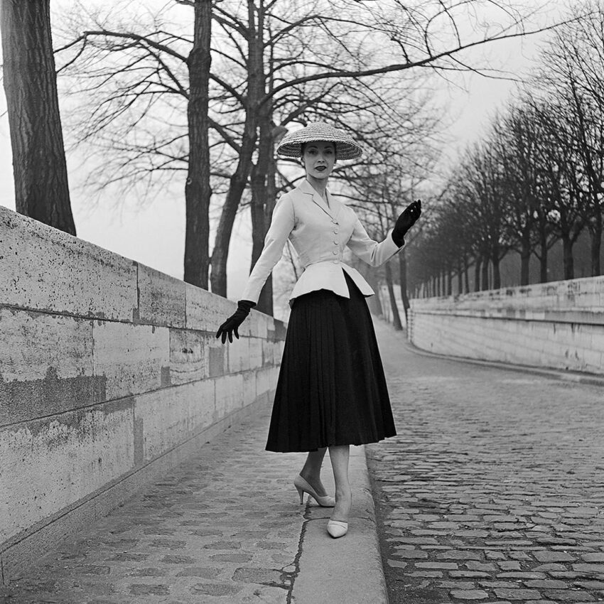 Vintage fashion model in a tailored jacket, full skirt, and hat, posing on a cobblestone path lined with bare trees.
