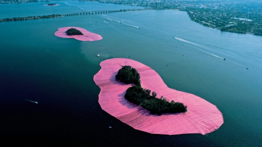 Aerial view of islands surrounded by vibrant pink fabric on a blue ocean, with distant buildings and boats.
