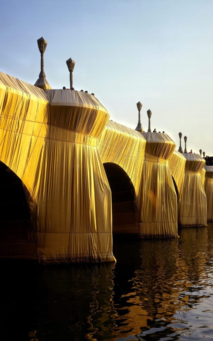 Golden draped bridge structures over water with reflections, set against a clear sky background.