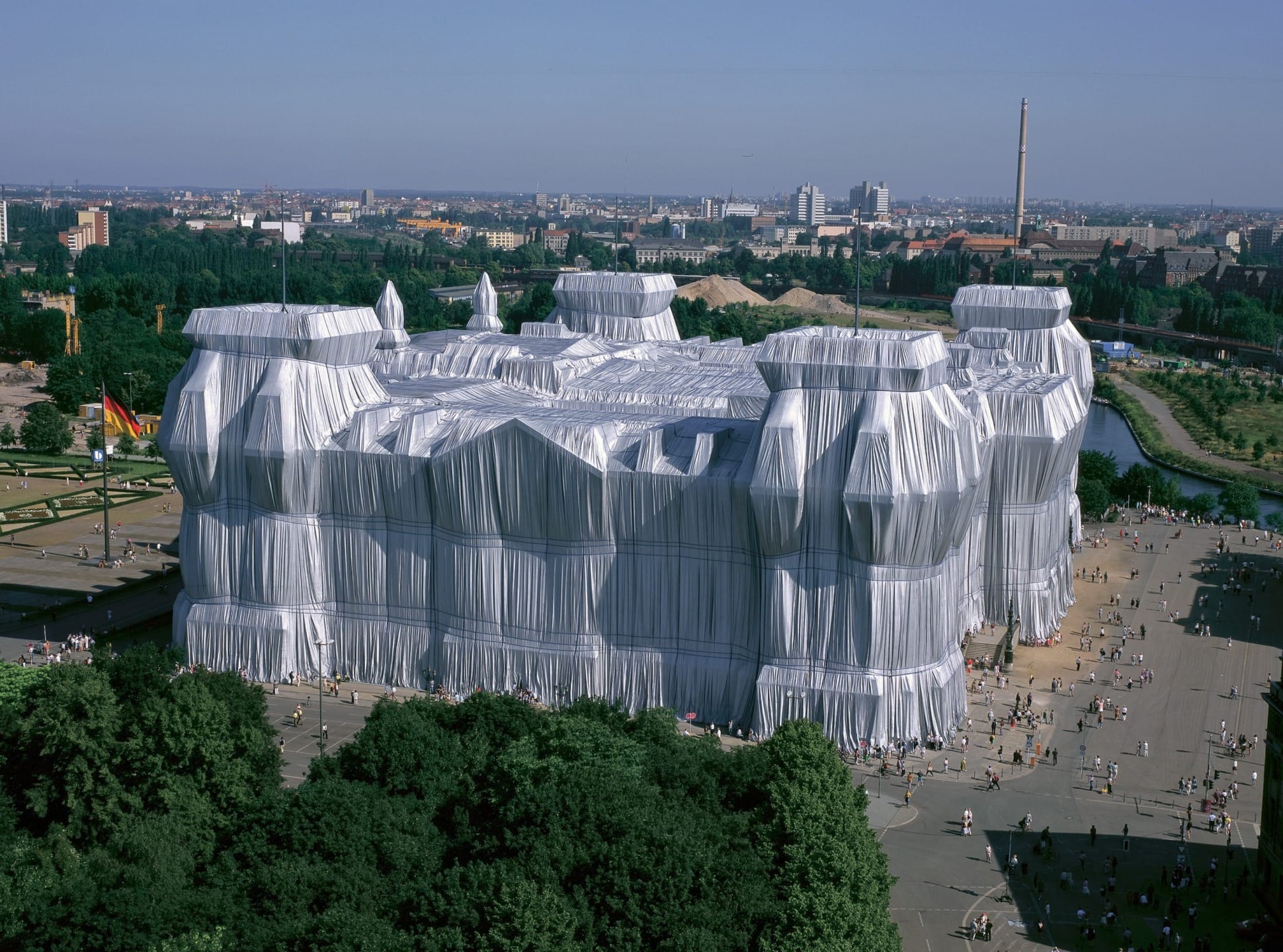Historic building wrapped in white fabric with a bustling crowd around, set against an urban and green landscape.