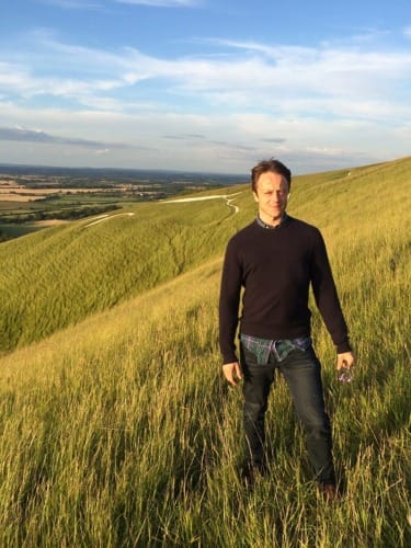 Person standing on a grassy hillside with a scenic view of fields and a winding path under a blue sky.