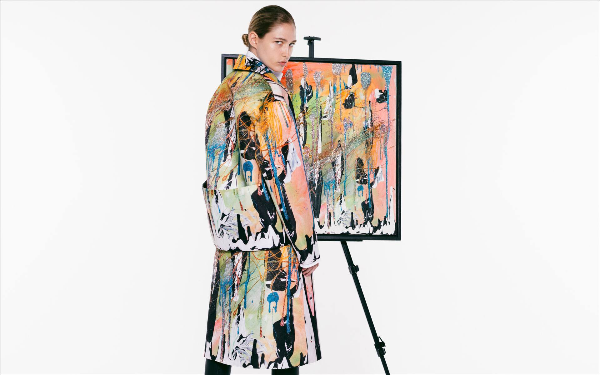 Person wearing a colorful coat stands next to an abstract painting on an easel against a plain background.