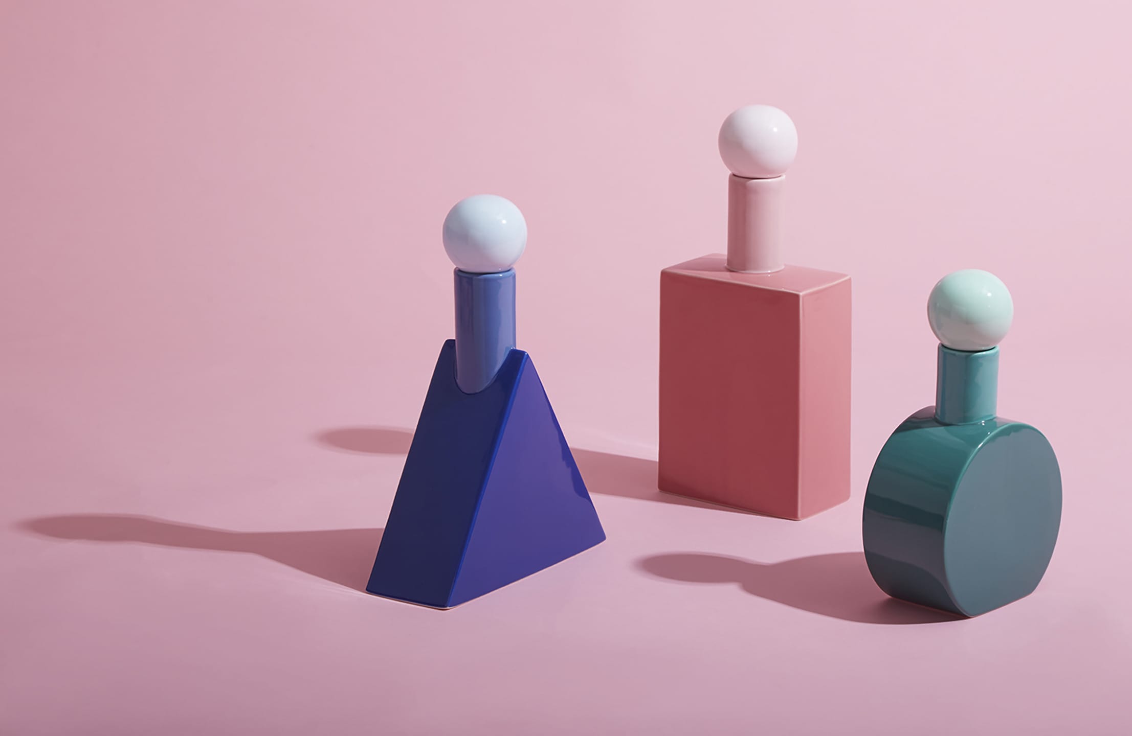 Colorful geometric perfume bottles on a pink background with soft lighting and long shadows.
