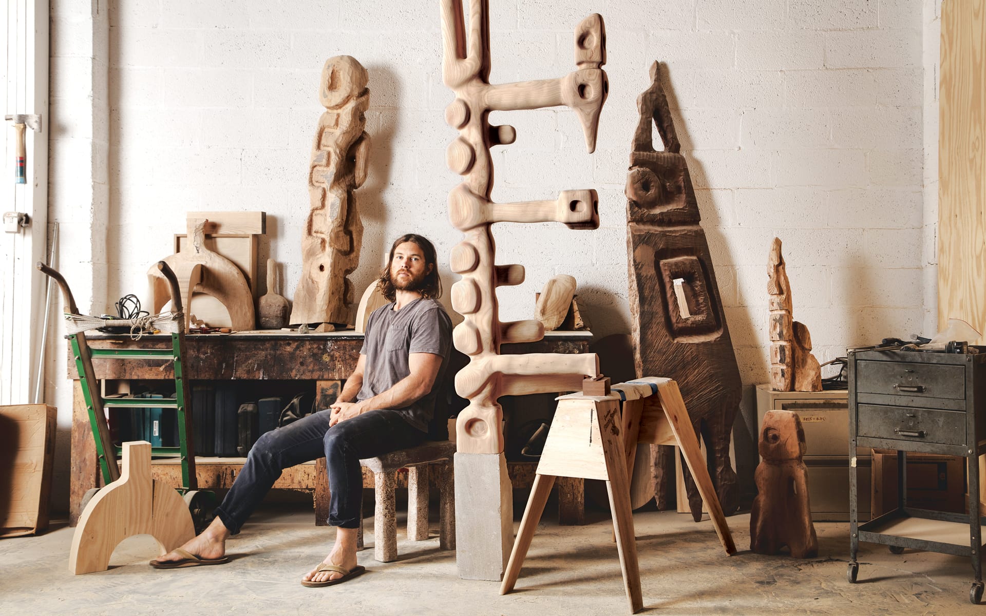 Man sitting in a workshop surrounded by abstract wooden sculptures and tools, wearing a casual outfit and sandals.