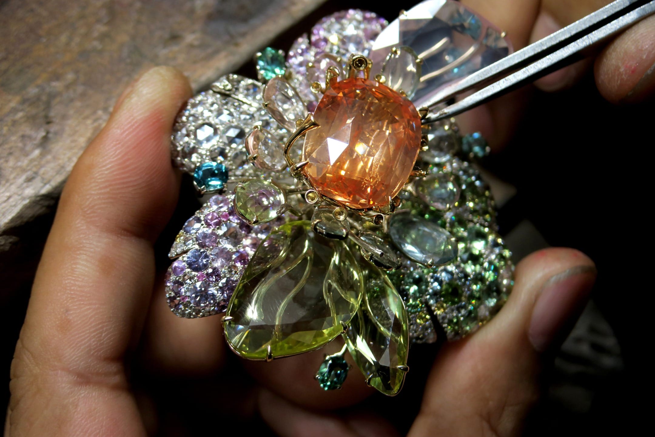 Close-up of hands crafting an ornate jeweled brooch with colorful gemstones and a central orange stone.