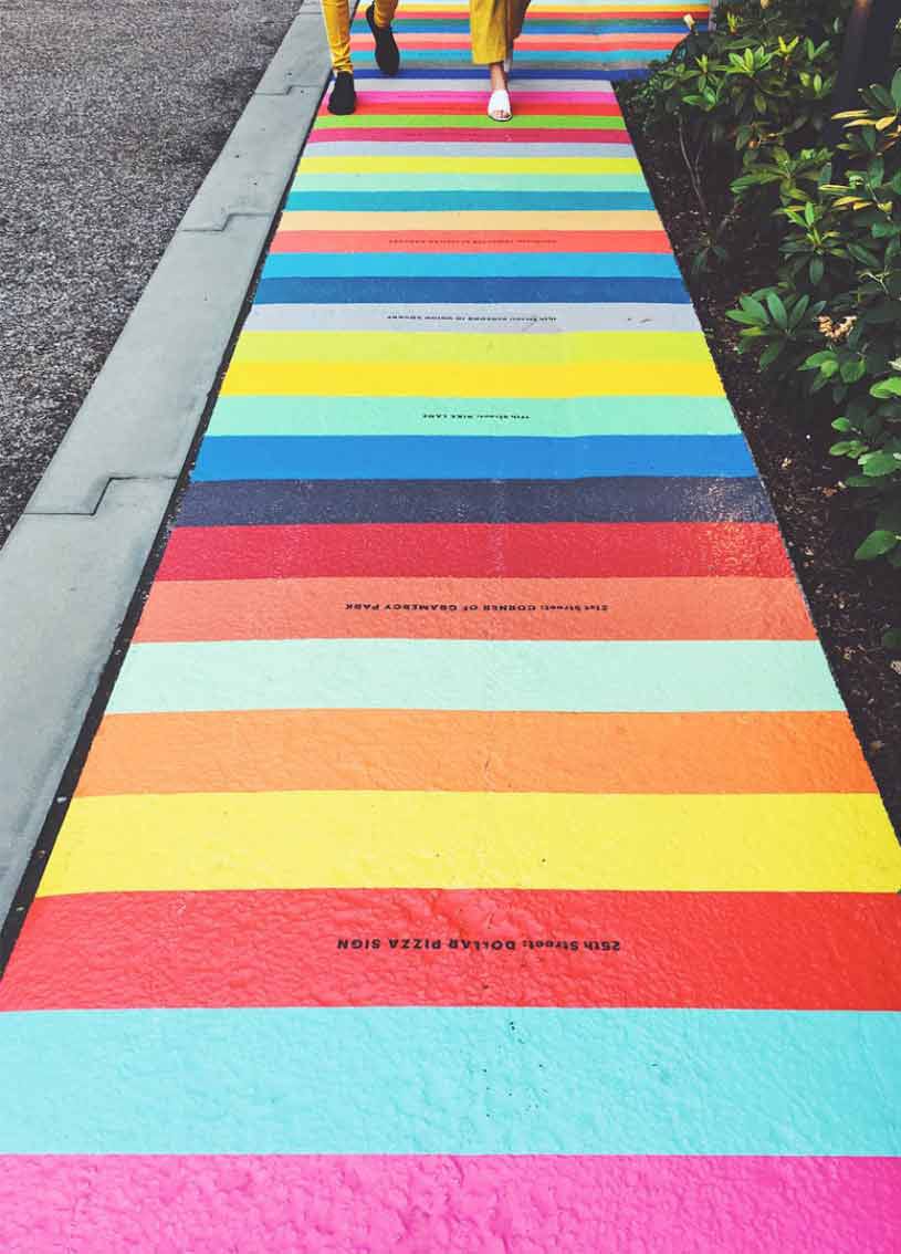 Colorful striped sidewalk with people walking on it, surrounded by green plants and a gravel path beside it.