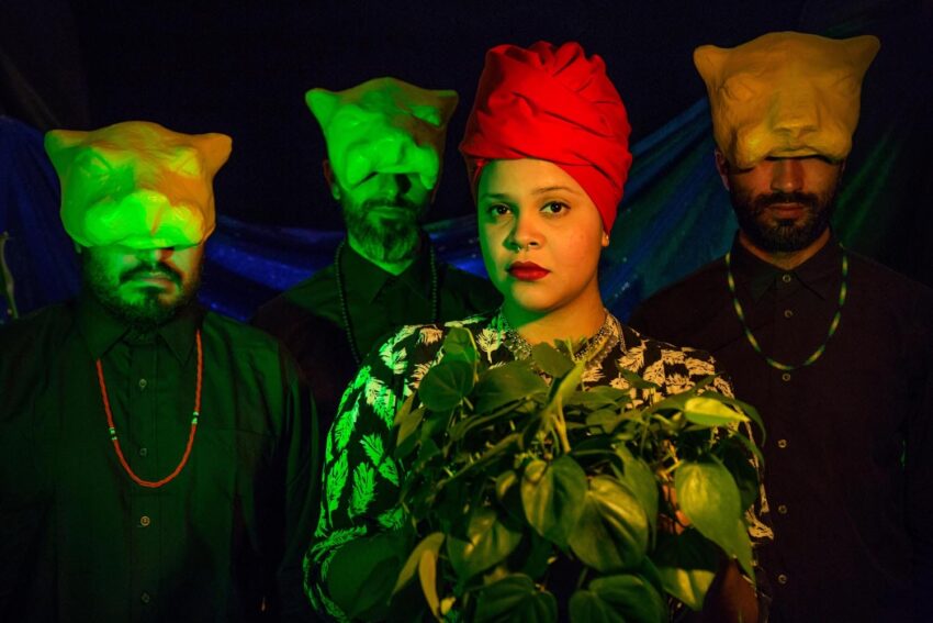 Person with red headwrap holds greenery, flanked by three people wearing green-lit animal masks, dramatic lighting and colors.