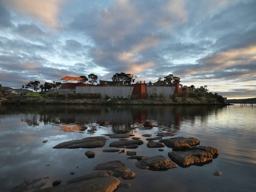 Modern building with metal and wood design reflected in calm water under a dramatic sky with scattered clouds.