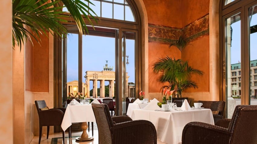 Elegant restaurant with a view of a famous city gate, featuring wicker chairs, white tablecloths, and lush indoor plants.