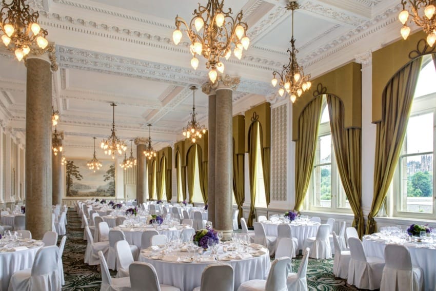 Elegant banquet hall with round tables, white chairs, hanging chandeliers, tall columns, and large windows with green curtains.