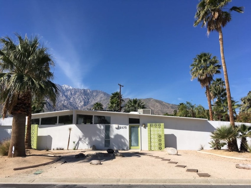 Mid-century modern house with green doors surrounded by palm trees and mountains under a clear blue sky.