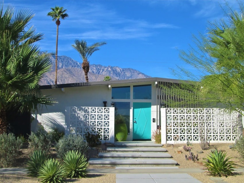 Mid-century modern house with turquoise door, surrounded by desert plants and mountains in the background under a blue sky.