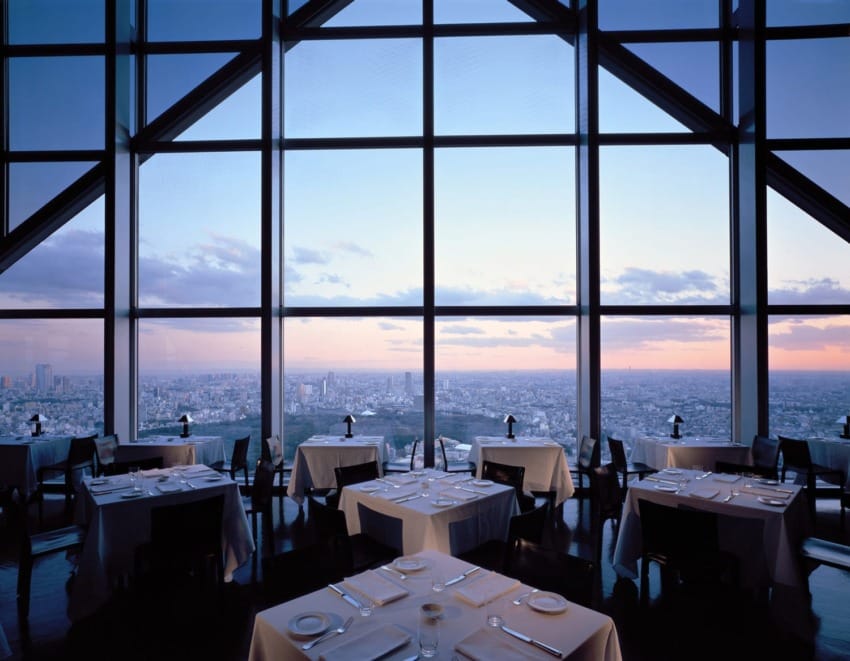 High-rise restaurant interior with large windows offering a panoramic city view at sunset, featuring neatly arranged tables.