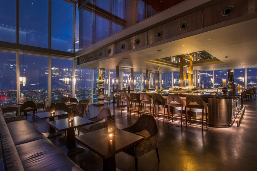 Rooftop bar interior at night with cityscape views, featuring cozy seating, tables, and a well-lit counter.