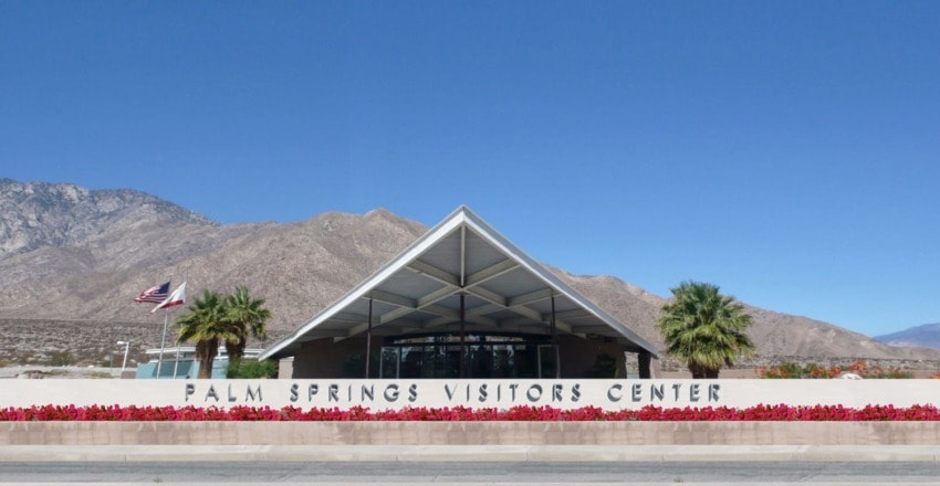 Palm Springs Visitors Center with a modern triangular roof, mountains in the background, flags waving, and red flowers in front.