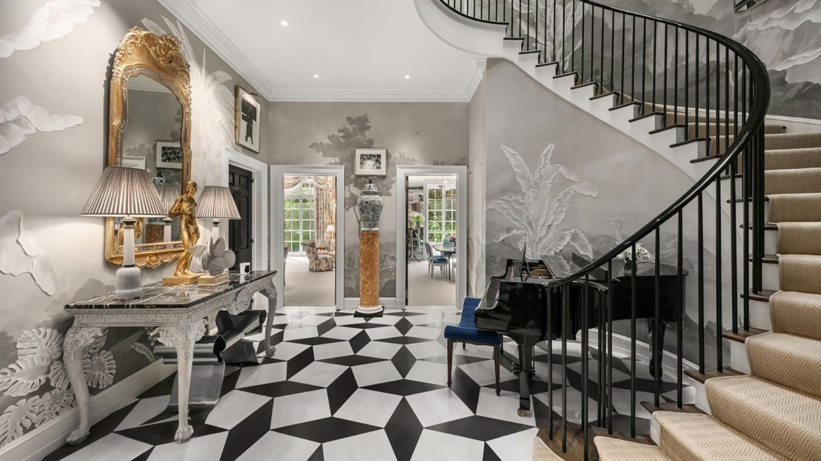 Elegant foyer with checkered floor, ornate mirror, and curved staircase with black railings, leading to a spacious room.