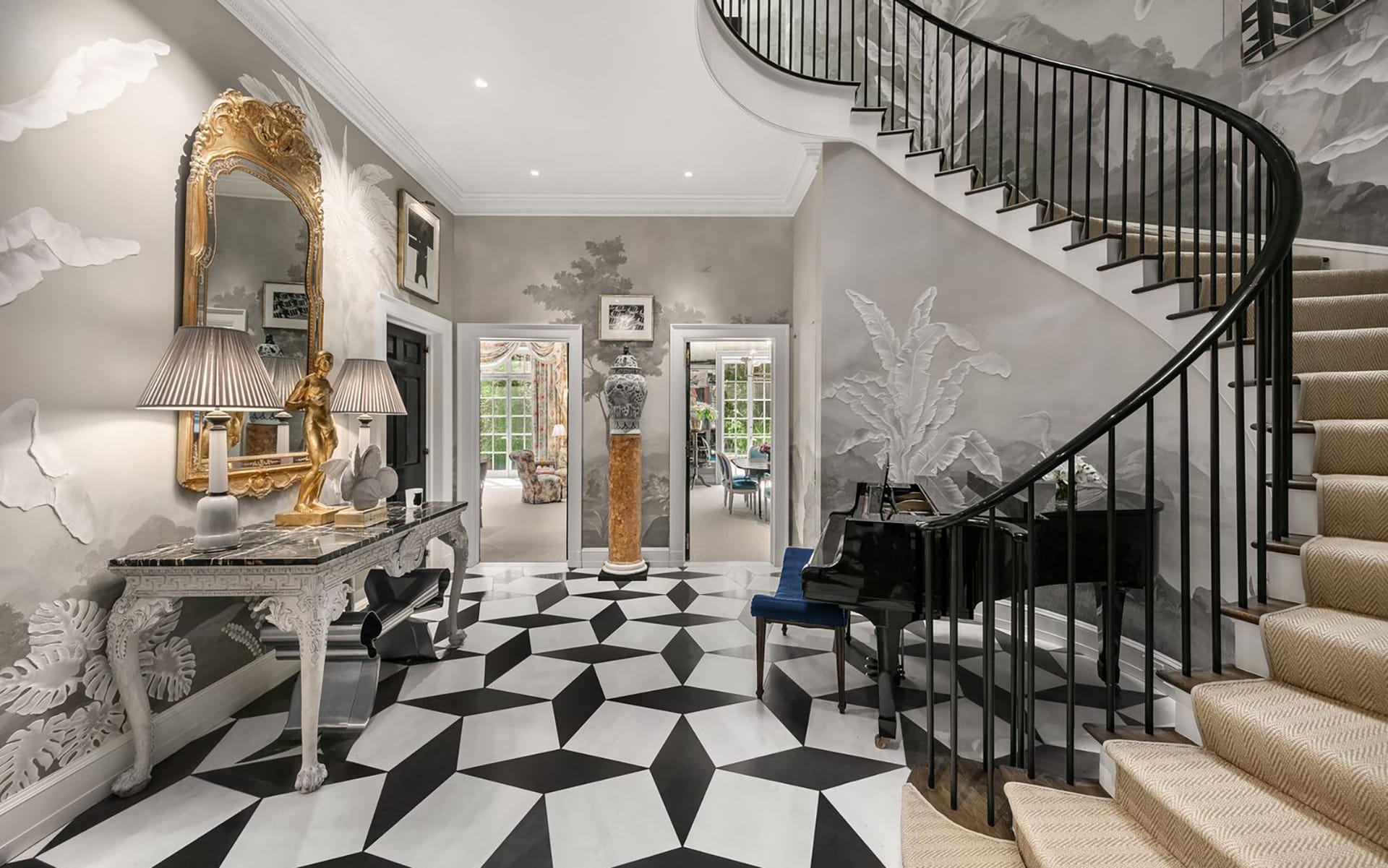 Elegant foyer with checkered floor, ornate mirror, and curved staircase with black railings, leading to a spacious room.
