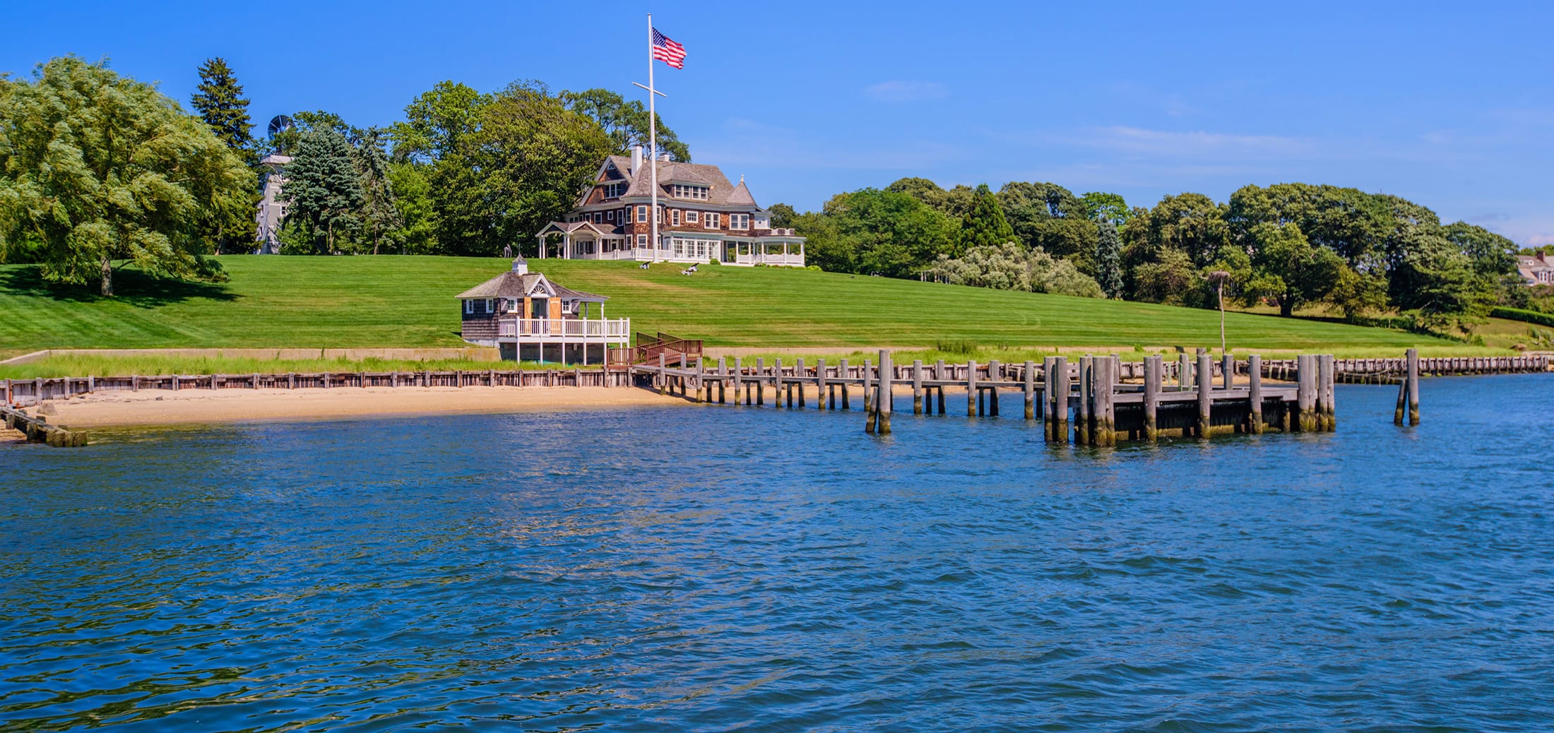 House with American flag on a hill, surrounded by greenery, overlooking a wooden dock and calm blue waters.