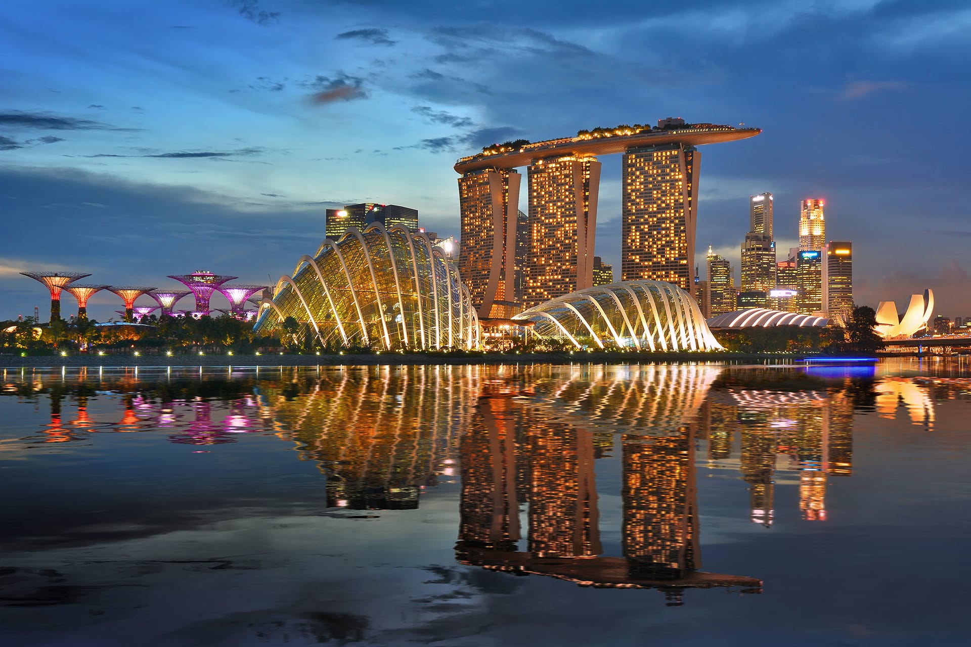 Singapore skyline at dusk with iconic Marina Bay Sands, Supertree Grove, and city lights reflecting on the water.