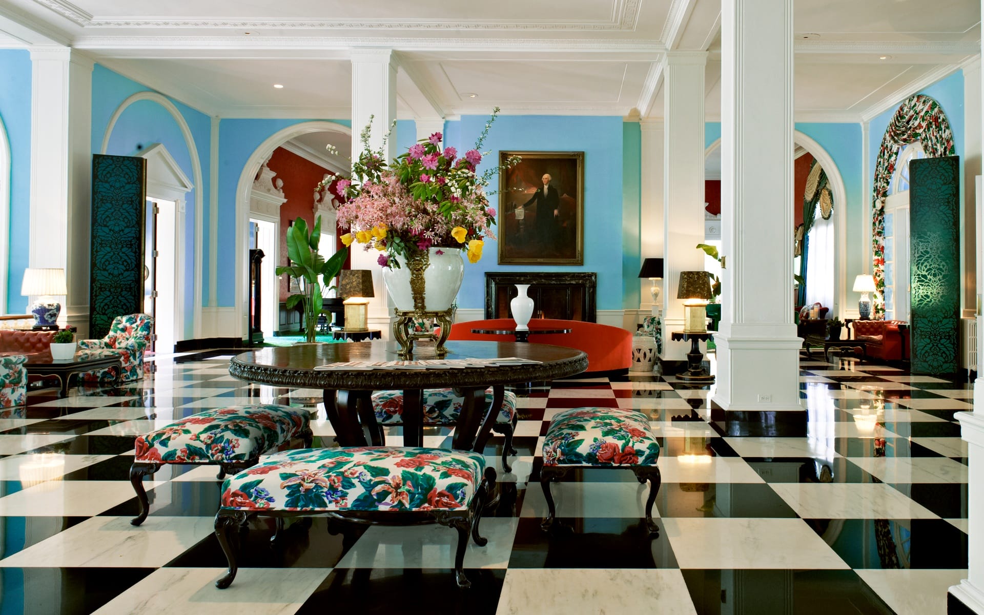 Elegant hotel lobby with checkered floor, floral furniture, large round table, colorful flowers, and blue walls.