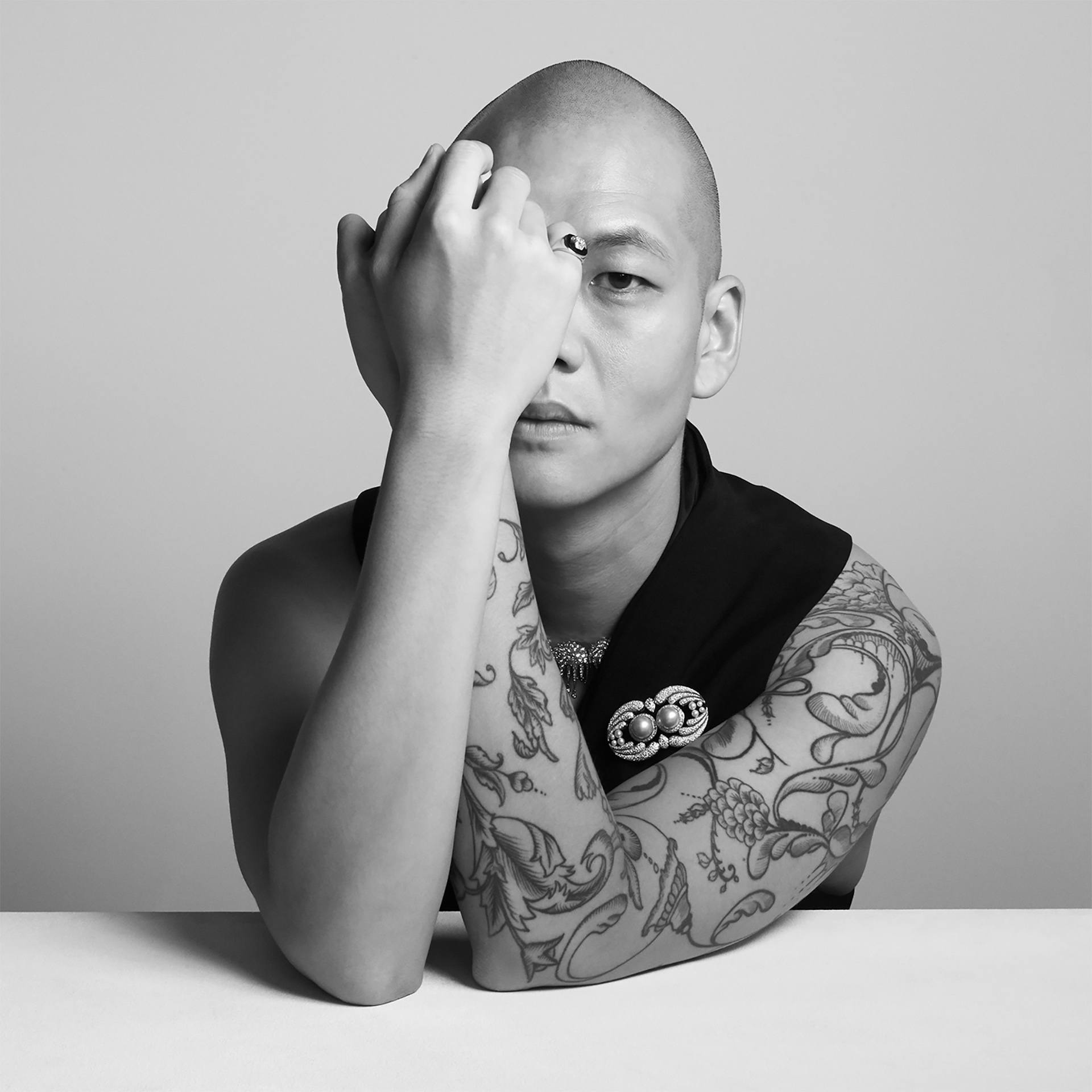 Person with tattoos, resting their head on their right hand, gazing directly into the camera, wearing a black sleeveless top.