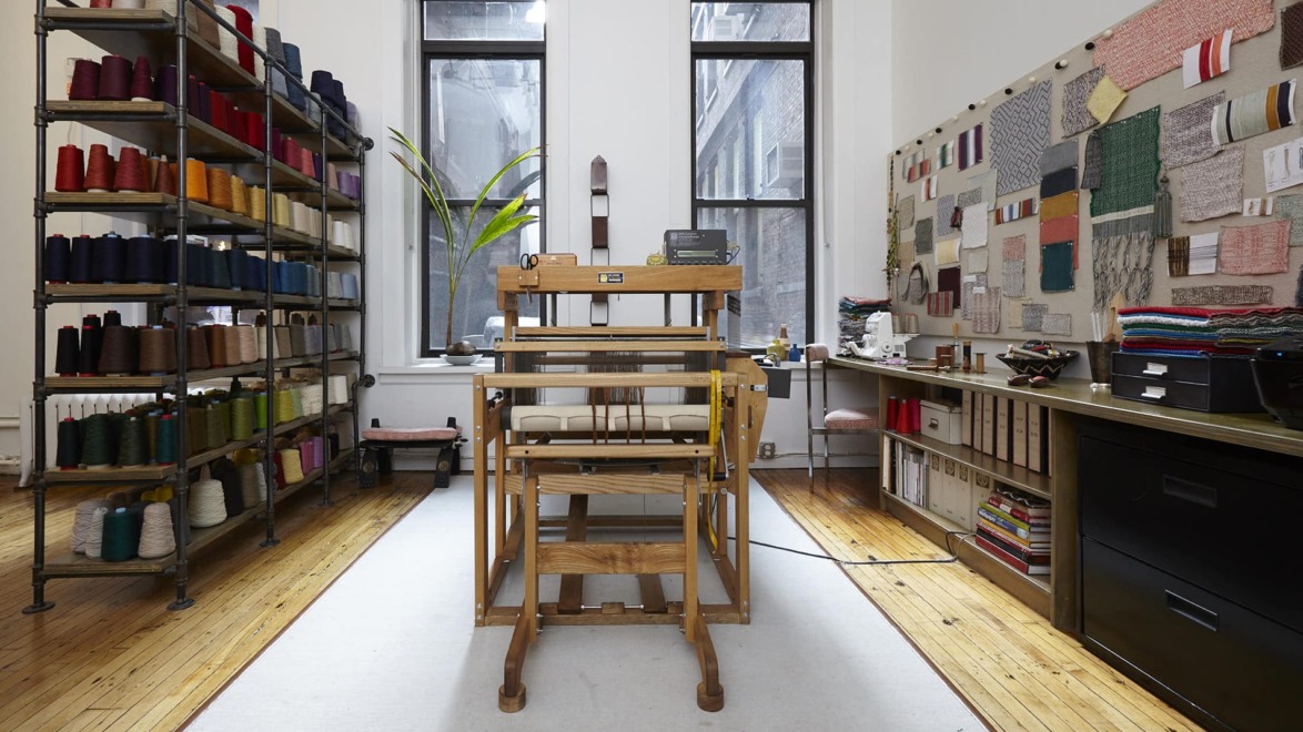 Wooden loom in a bright textile studio with colorful thread spools on shelves and fabric swatches on the wall.