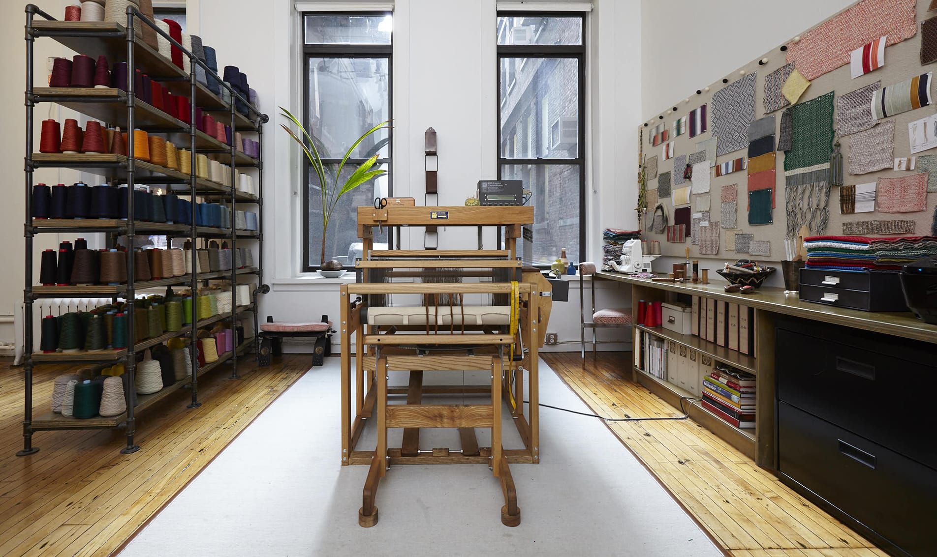 Wooden loom in a bright textile studio with colorful thread spools on shelves and fabric swatches on the wall.