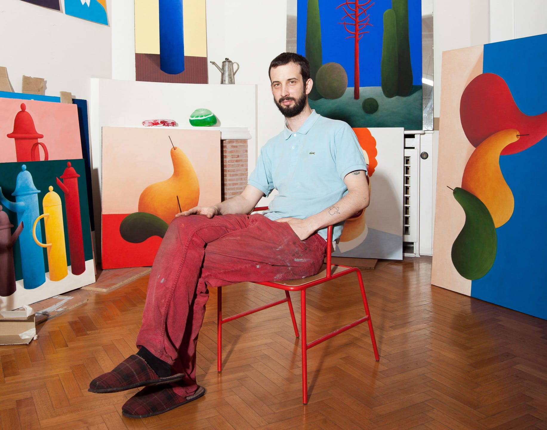 Artist seated in studio surrounded by colorful abstract paintings on canvas, wearing red pants and slippers.
