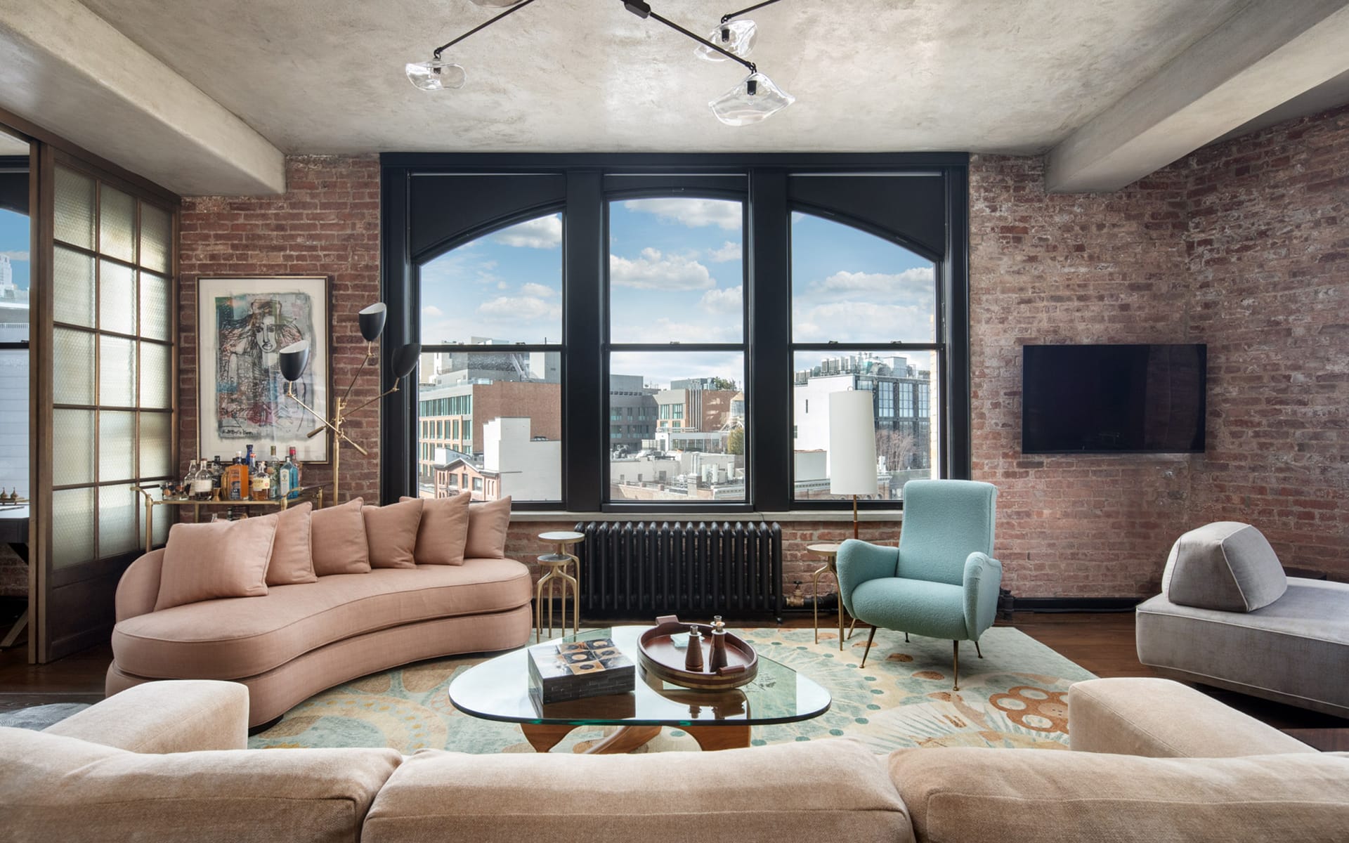 Loft-style living room with large windows, exposed brick walls, modern furniture, and cityscape view.