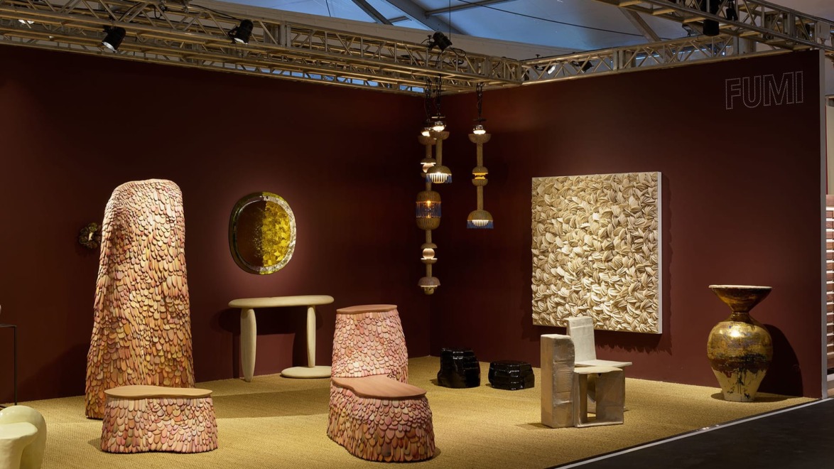 Art gallery booth with abstract wooden furniture, sculptures, and wall art in a warmly lit exhibition space.
