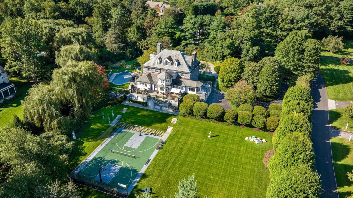 Aerial view of a large mansion with a circular driveway, lush gardens, a swimming pool, and a sports court in a wooded area.