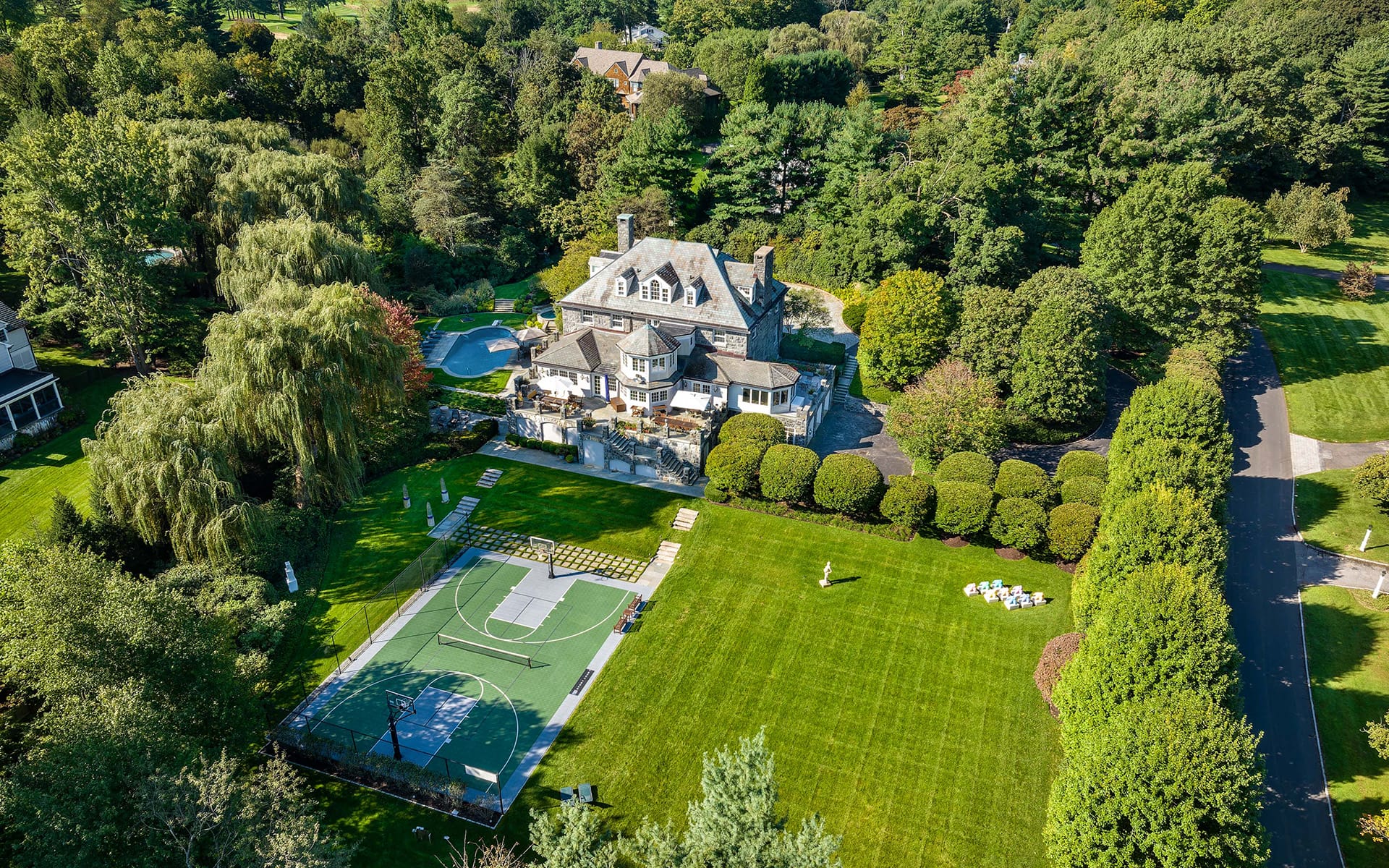 Aerial view of a large mansion with a circular driveway, lush gardens, a swimming pool, and a sports court in a wooded area.