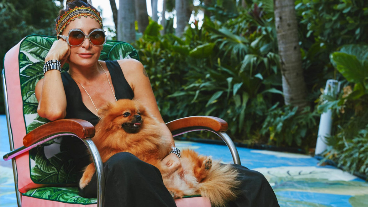 Woman sitting in a tropical garden chair holding a fluffy dog, wearing sunglasses and a black outfit, surrounded by lush greenery.