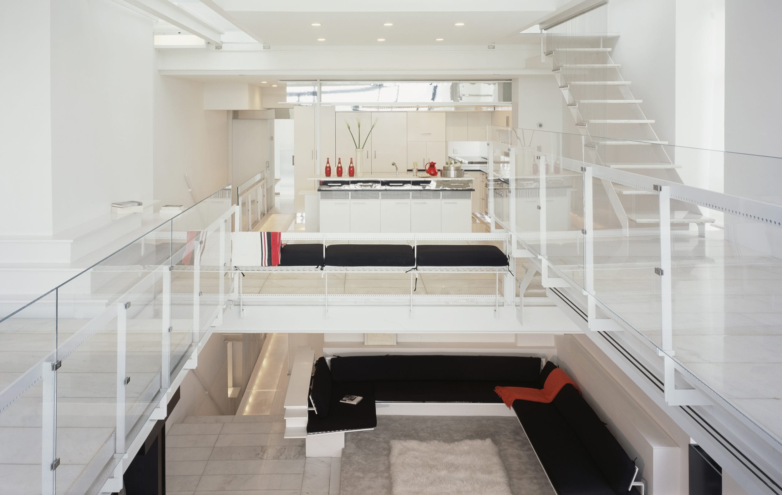 Modern, open-concept white living space with glass railings, minimalist furniture, and upper-level view of kitchen area.