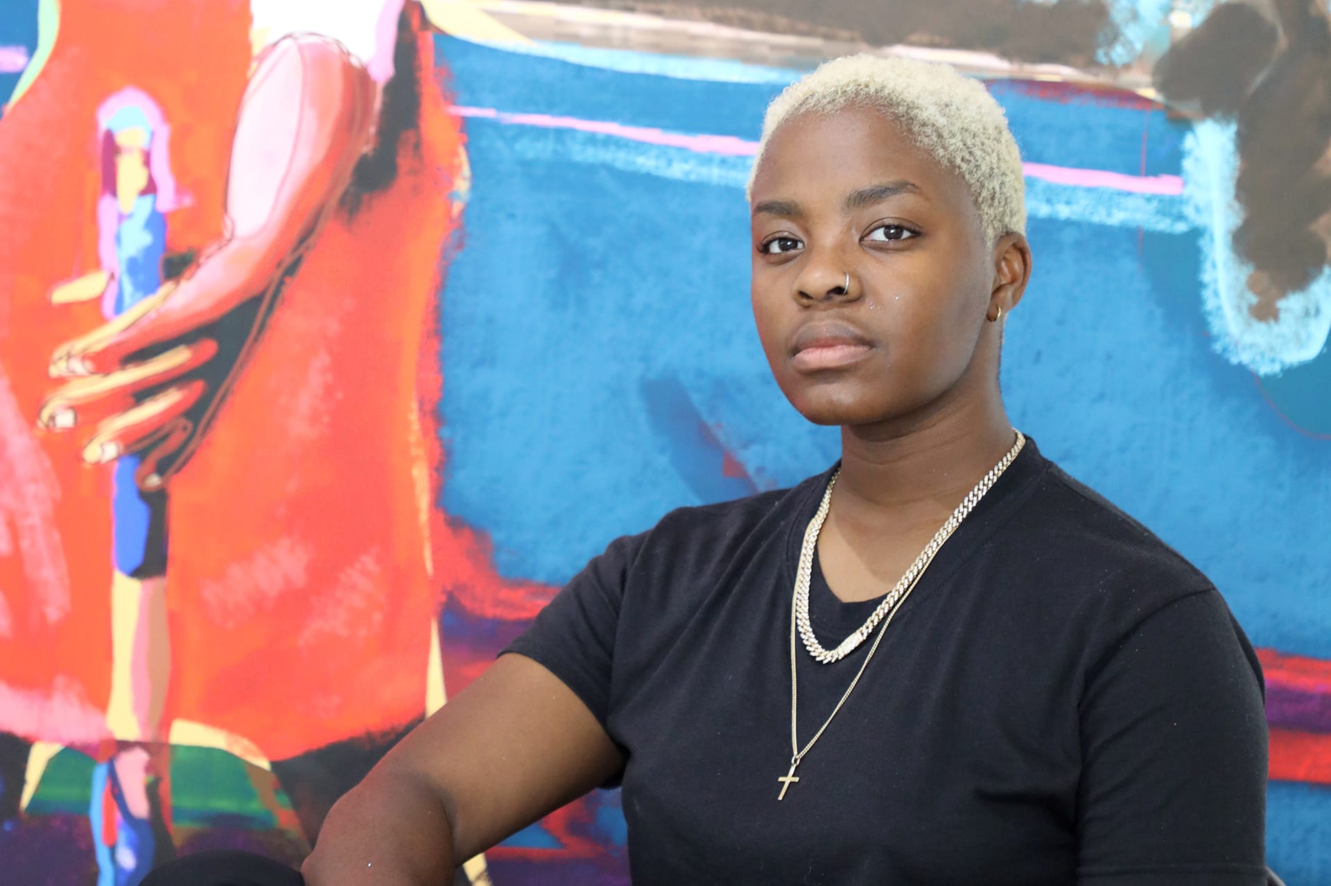 Person with short hair wearing black t-shirt and necklace, sitting in front of a colorful abstract painting background.