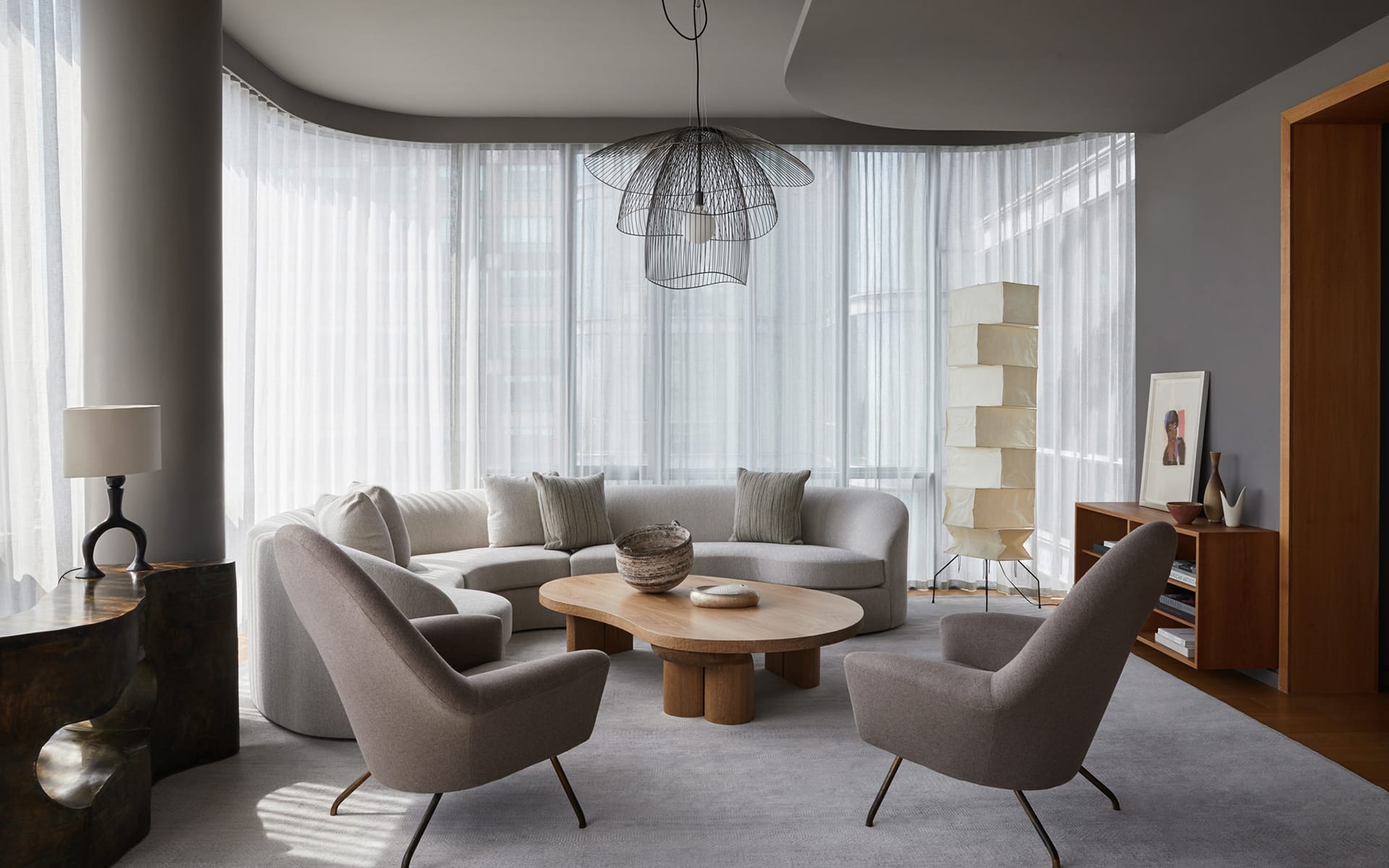 Modern living room with large windows, curved sofa, two armchairs, round coffee table, and contemporary lighting fixture.